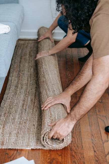 A person is rolling out a beige and brown patterned carpet onto a wooden floor in a living room, with another individual helping to smooth it out. The room features hardwood flooring, and the carpet a