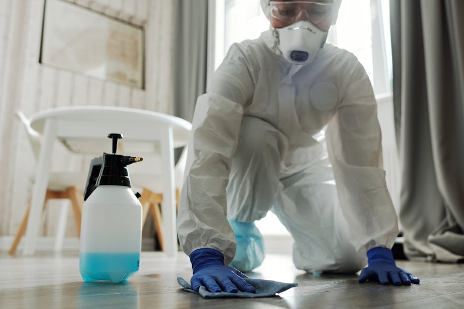 A professional cleaner wearing protective gear including a white suit, face mask, goggles, and blue gloves is using a high-pressure steam cleaning tool to sanitize a surface inside a commercial or res